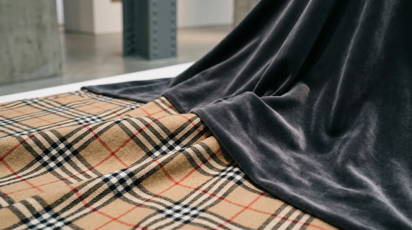 Tartan classique partiellement recouvert d'un velours moderne sombre, métaphore visuelle du patrimoine remis en couverture après une parenthèse minimaliste